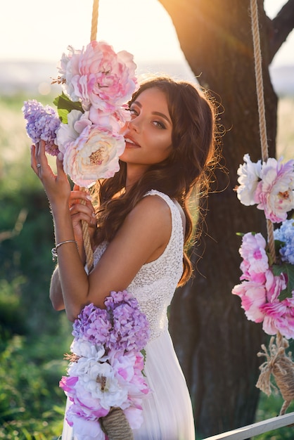 Sinnliches süßes Mädchen in einem weißen Kleid nahe einer hölzernen Schaukel verziert mit zarten farbigen Blumen im Sonnenuntergang.
