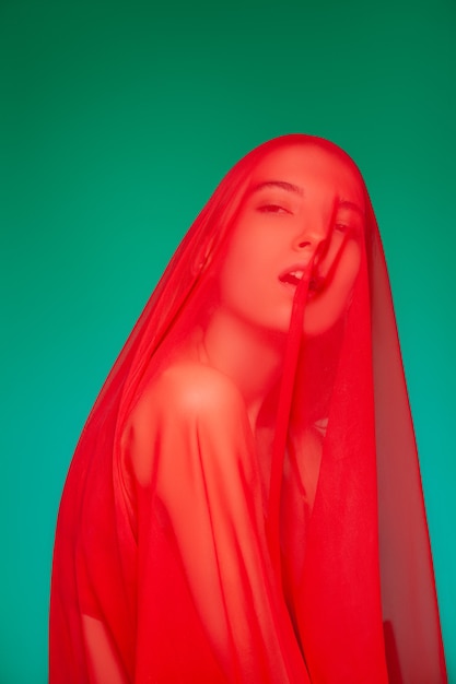 Sinnliches weibliches Modell im roten durchscheinenden Umhang, der Gesicht und Körper bedeckt und mit geschlossenen Augen vor grünem Hintergrund träumt
