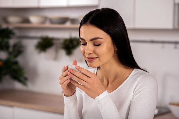 Foto sinta-se uma linda mulher encantada bebendo chá e apreciando seu sabor na cozinha