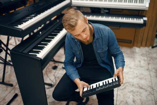 Sintetizador de compra de músico joven barbudo en la tienda de música. Surtido en la tienda de instrumentos musicales, músico masculino elegir teclado