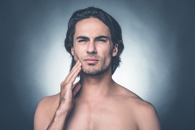 Sintiendo ese terrible dolor de muelas. Retrato de hombre joven sin camisa que expresa negatividad mientras toca la mejilla y de pie contra el fondo gris