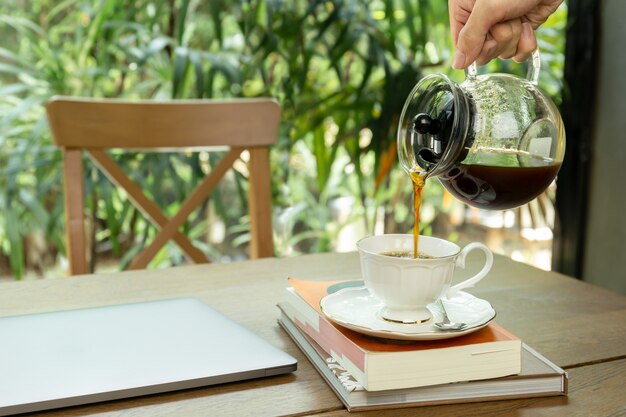 Sirva el café de colada en una taza con el ordenador portátil en la tabla.