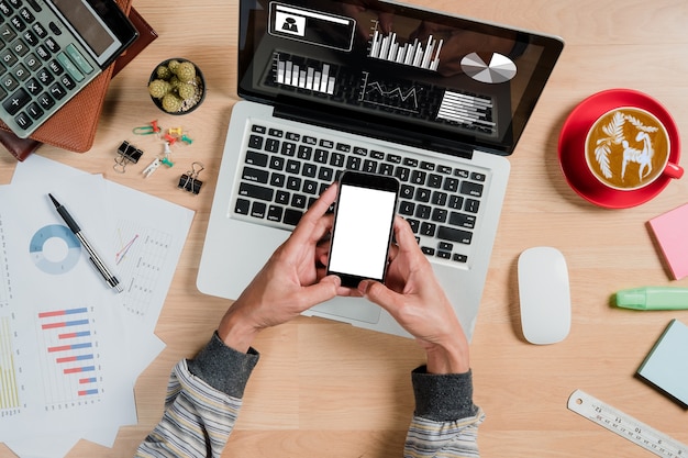 Sirva las manos que sostienen la pantalla en blanco un smartphone en el escritorio de trabajo.