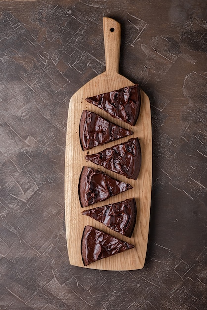 Sirviendo brownie triangular con crema de ganache en una tabla de madera para servir. Vista superior, endecha plana, fondo oscuro.