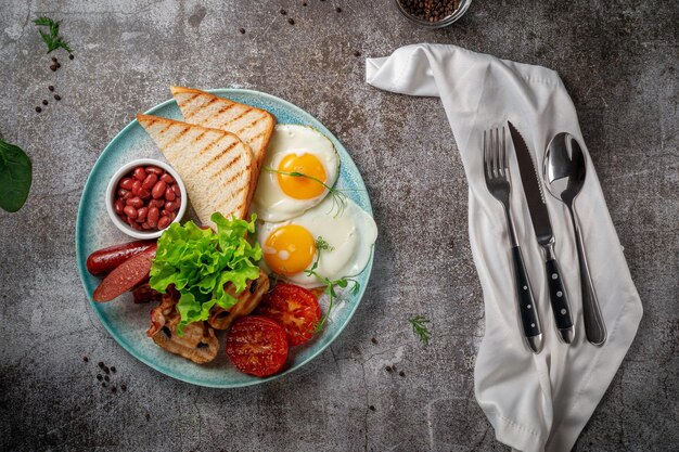 Sirviendo un plato de la carta del restaurante. Delicioso y saludable desayuno con huevos fritos, frijoles, salchichas, verduras asadas, tomates y champiñones con ensalada en un plato contra una mesa de piedra gris