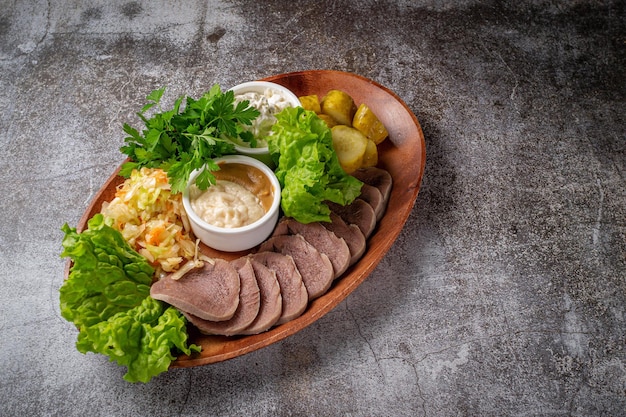 Sirviendo un plato de la carta del restaurante. Lengua de ternera en rodajas con encurtidos, repollo, salsas y hierbas en un plato contra una mesa de piedra gris, un delicioso aperitivo