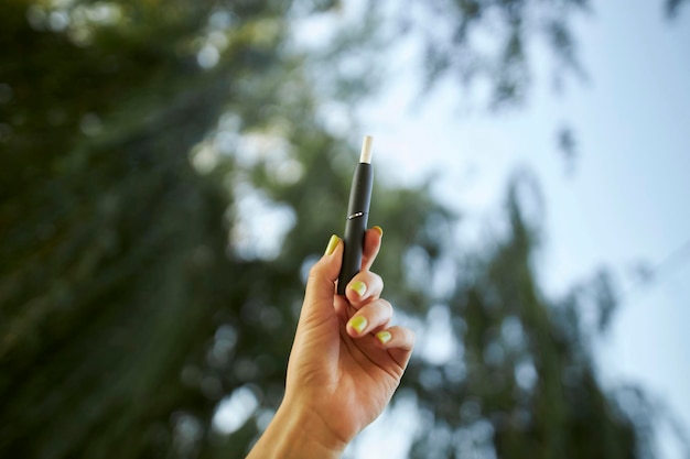 Sistema de calentamiento de tabaco IQOS sobre fondo de verano. Mano de mujer sosteniendo un cigarrillo electrónico.