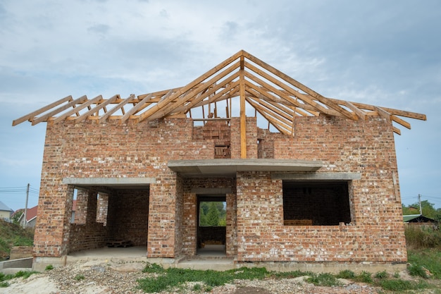 Foto sitio de construcción con casa de ladrillo sin terminar con marco de techo de madera para el futuro techo en construcción.