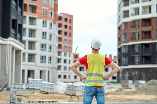 Foto sitio de construcción. vista posterior del hombre en casco protector sosteniendo sus manos en el cinturón mirando casas en construcción en sitio de construcción