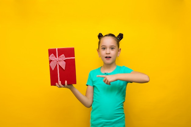 La situación feliz de la niña aislada sobre fondo amarillo señala en la caja de regalo roja en sorpresa.