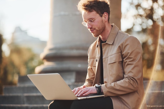 Sitzen auf der Treppe mit Laptop auf den Beinen Eleganter junger Mann in formeller, eleganter Kleidung im Freien in der Stadt