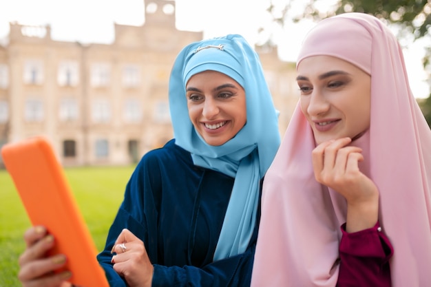 Sitzen in der Nähe von Freund. Student mit blauem Hijab, der ein orangefarbenes Tablet hält, das in der Nähe eines Freundes mit Laptop sitzt