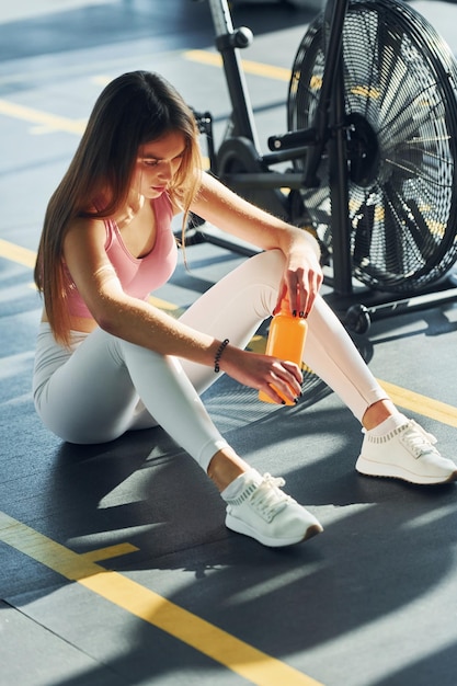 Sitzen mit Flasche Wasser Schöne junge Frau mit schlankem Körpertyp ist im Fitnessstudio