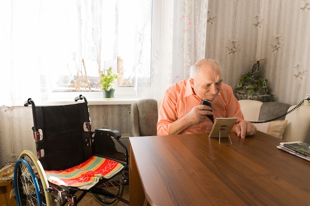 Sitzender älterer Mann beim Rasieren von Bärten mit Elektrorasierer im Wohnzimmer mit seinem Rollstuhl auf der Seite.