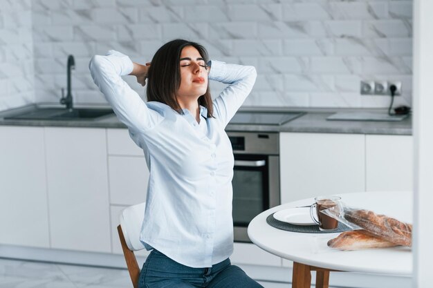 Sitzt am Tisch mit Morgenmahlzeit Junge Frau ist tagsüber drinnen im Zimmer eines modernen Hauses