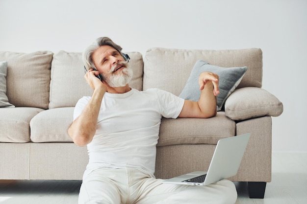 Sitzt auf dem Boden in der Nähe des Sofas mit Laptop und hört Musik Senior stilvoller moderner Mann mit grauem Haar und Bart drinnen