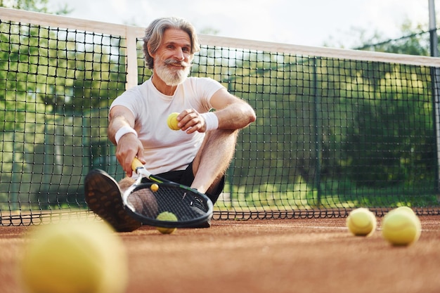 Sitzt auf dem Boden und macht Pause Älterer, moderner, stilvoller Mann mit Schläger im Freien auf dem Tennisplatz tagsüber