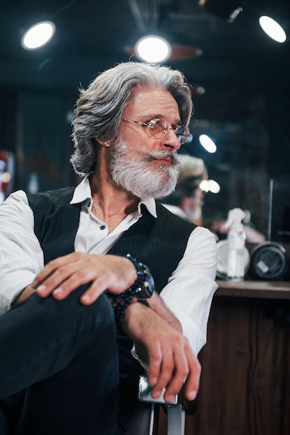 Sitzt auf dem Stuhl im Barbershop Stilvoller moderner Senior mit grauem Haar und Bart ist drinnen