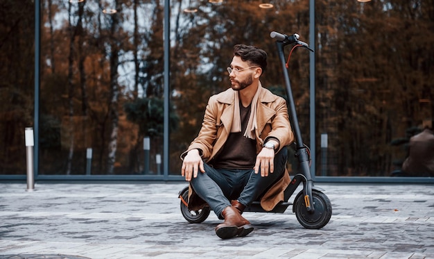 Sitzt auf Elektroroller Junges männliches Model in modischer Kleidung ist tagsüber draußen in der Stadt