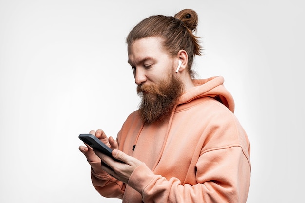 Skandinavischer gutaussehender ernster Mann, der das Telefon mit drahtlosen Kopfhörern betrachtet, die auf grauem Hintergrund in rosafarbenem orangefarbenem Sweatshirt isoliert sind. Ruhiger Kerl mit Ingwerfrisur und Bart. Moderne digitale Technologien