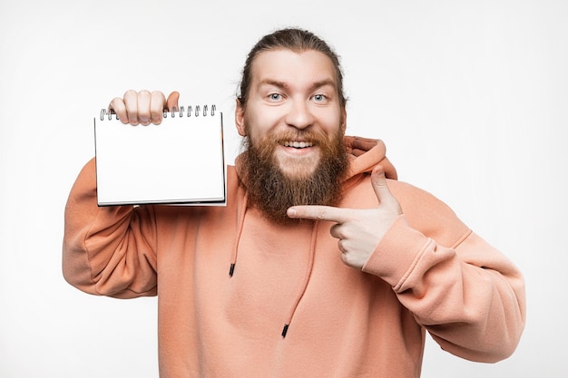 Foto skandinavischer gutaussehender glücklicher mann mit notizblock und verweisen auf papiermockup isoliert auf grauem hintergrund kerl mit ingwerfrisur und bart in einem sweatshirt kerl mit notebook-platz für werbung