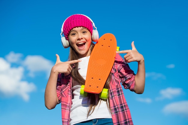 Skate em que você pode confiar. Garota feliz apontando para o tabuleiro de um centavo. Pequena patinadora com gesto apontando no céu azul. Apontando para publicidade. Marketing e promoção. Bom para transporte, espaço de cópia.