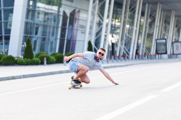 Skateboarder mit Sonnenbrille fährt Longboard auf asphaltierter Straße vor dem modernen Gebäude in der Stadt mit Trickfahrt in niedriger Position