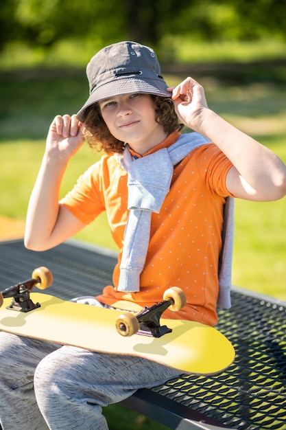 Skater adolescente con la tabla de rodillas sentado en el banco de acero perforado y enderezándose el sombrero