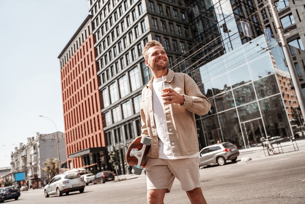 Skater joven rubio guapo sosteniendo longboard en el hombro y una taza de café para llevar mirando alegre sonriendo mientras camina en el fondo del edificio urbano. Viste un atuendo casual de mezclilla beige.