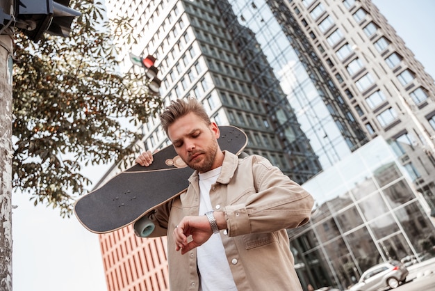 Skater joven rubio guapo sosteniendo longboard en el hombro a toda prisa mirando serio en el reloj de pulsera. Fondo de edificio urbano en movimiento. Viste camisa de mezclilla beige. Ocio al aire libre. Viajar diariamente