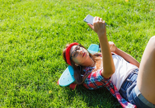 Skater de mujer con estilo divertido con patín tumbado en la hierba verde y escuchando música en auriculares en un día soleado