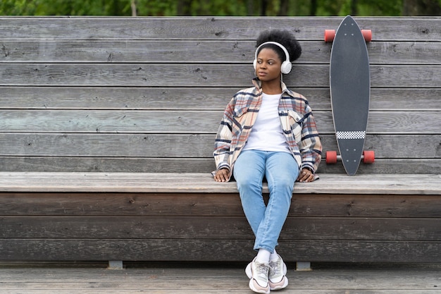 Skater de mujer negra relajado escuchar música sentarse en el parque con concepto de estilo de vida urbano de tablero largo