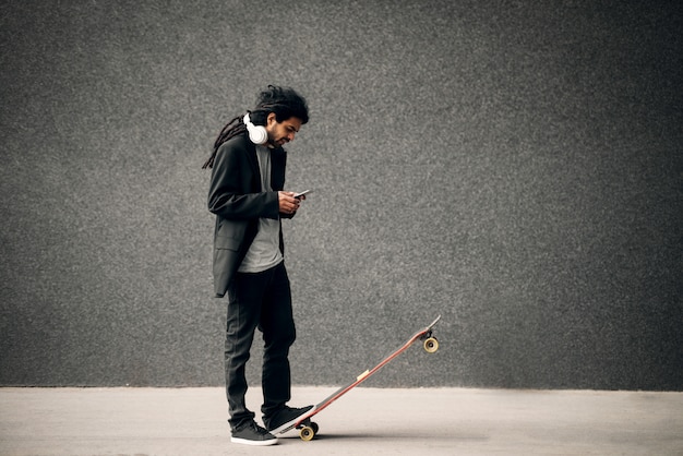 Skatista de hipster elegante jovem dreadlocks com fones de ouvido, olhando para o telefone inteligente em pé na prancha longa.