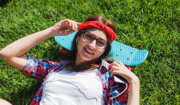 Skatista engraçada mulher elegante com skate deitado na grama verde e ouvir música em fones de ouvido em dia ensolarado