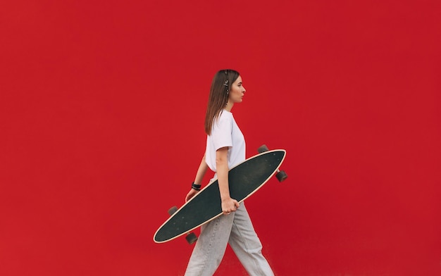 Skatista jovem confiante em roupa casual está andando na frente de uma parede vermelha isolada