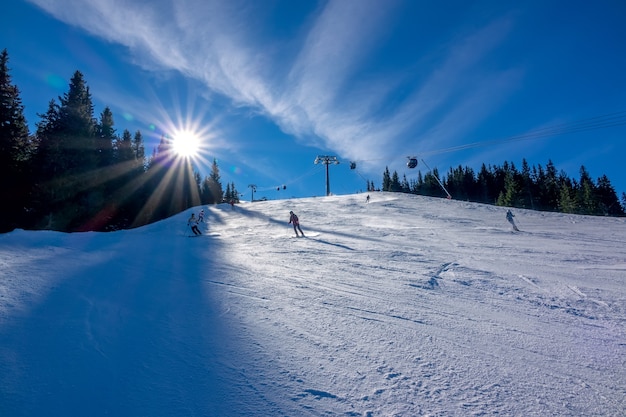 Skifahrer gehen einen breiten Hang hinunter. Bäume, Sonne und Skilift