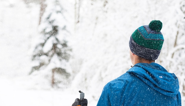 Skifahrer in Windjacke und Hut mit Pompon mit Skistöcken in den Händen mit dem Rücken auf einem verschneiten Wald. Langlauf im Winterwald, Outdoor-Sport, gesunder Lebensstil.