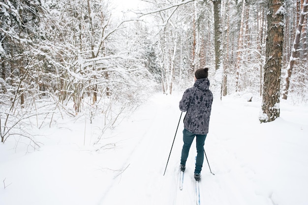 Skifahrer mit Hut mit Bommel mit Skistöcken in den Händen mit dem Rücken vor dem Hintergrund eines verschneiten Waldes Langlaufen im Winterwald im Freien Sport gesunder Lebensstil