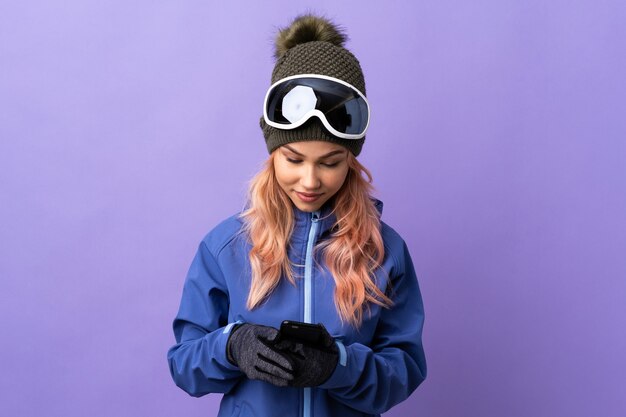 Skifahrer-Teenager-Mädchen mit Snowboardbrille über isolierter lila Wand, die eine Nachricht mit dem Handy sendet