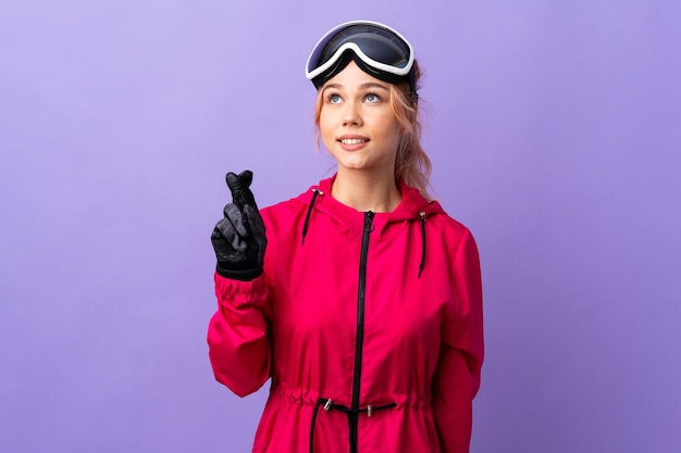 Skifahrer-Teenager-Mädchen mit Snowboardbrille über isolierter lila Wand mit den Fingern, die kreuzen und das Beste wünschen