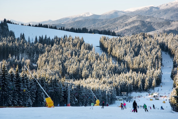 Skifahrer und Snowboarder bergab im Winterskigebiet vor dem Hintergrund von Skiliften, Wäldern, Hügeln am sonnigen Abend.