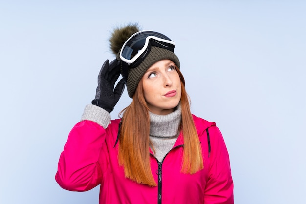 Skifahrerrothaarigefrau mit Snowboardinggläsern über der lokalisierten blauen Wand, die Zweifel hat und mit verwirren Gesichtsausdruck