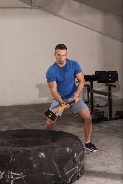 Sledgehammer Tire Golpea a un joven musculoso entrenando en el gimnasio con un martillo y un neumático de tractor