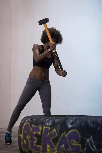 Sledgehammer Tire Golpea a una mujer afroamericana entrenando en el gimnasio con un martillo y un neumático de tractor