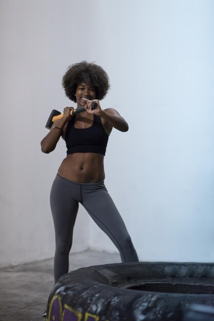 Sledgehammer Tire Golpea a una mujer afroamericana entrenando en el gimnasio con un martillo y un neumático de tractor