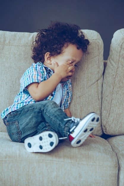 Sleepycutie. Pequeño bebé africano con sueño y frotándose los ojos mientras está sentado en el sofá en casa