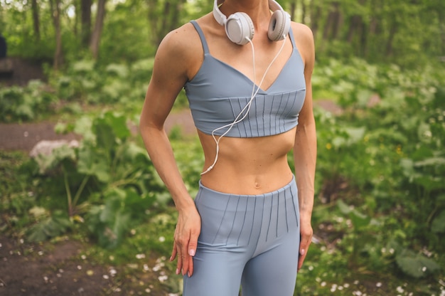 Slim Fit chica en ropa deportiva azul con grandes auriculares blancos haciendo deporte y fitness en el parque de verano afuera