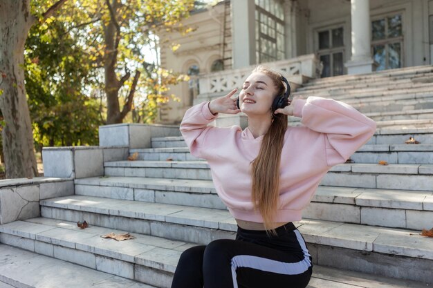 Slim fit mulher atraente sentada nos degraus e ouvindo música com fones de ouvido ao ar livre