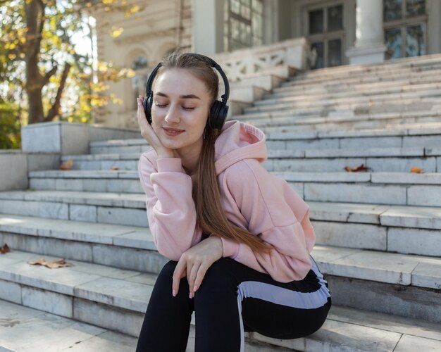 Slim fit mulher atraente sentada nos degraus e ouvindo música com fones de ouvido ao ar livre