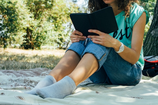 Slim garota de jeans e uma camiseta, lendo um livro em um parque, sentado em um cobertor